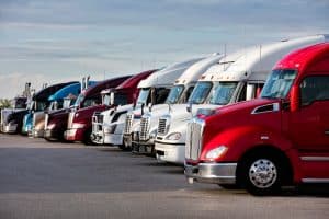 A row of semi-trucks parked in a sunny parking lot. While none of the trucks have visible truck windscreen sun shades, all the windshields have sun visors folded down to block the sun.