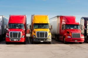 The image shows a row of three semi-trucks parked in a parking lot. There are sun visors attached to the inside of the windscreens, but these are built into the trucks themselves and are not removable sun shades.